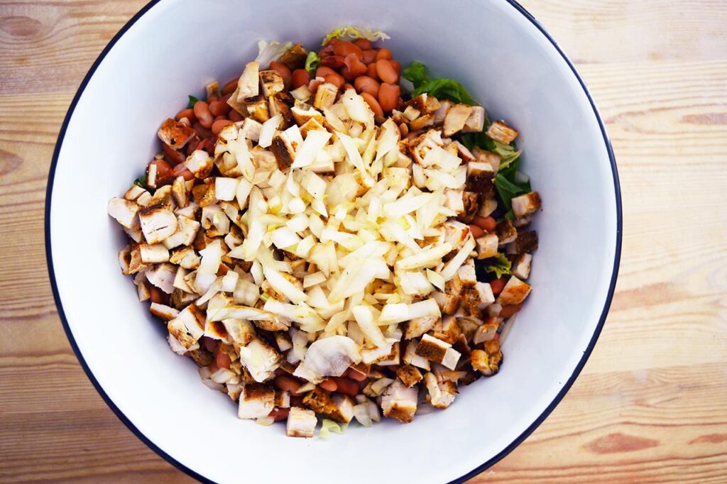 Chopped onions layered over chopped chicken, beans and lettuce in a mixing bowl.