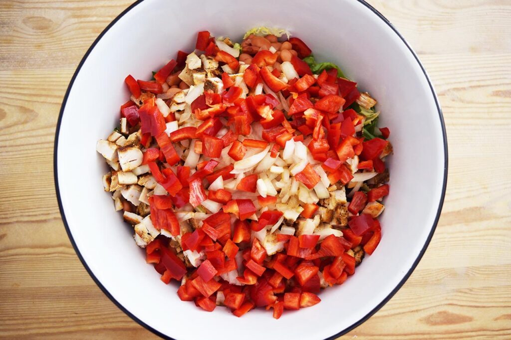 A layer of chopped red bell peppers added over the salad in a white mixing bowl.