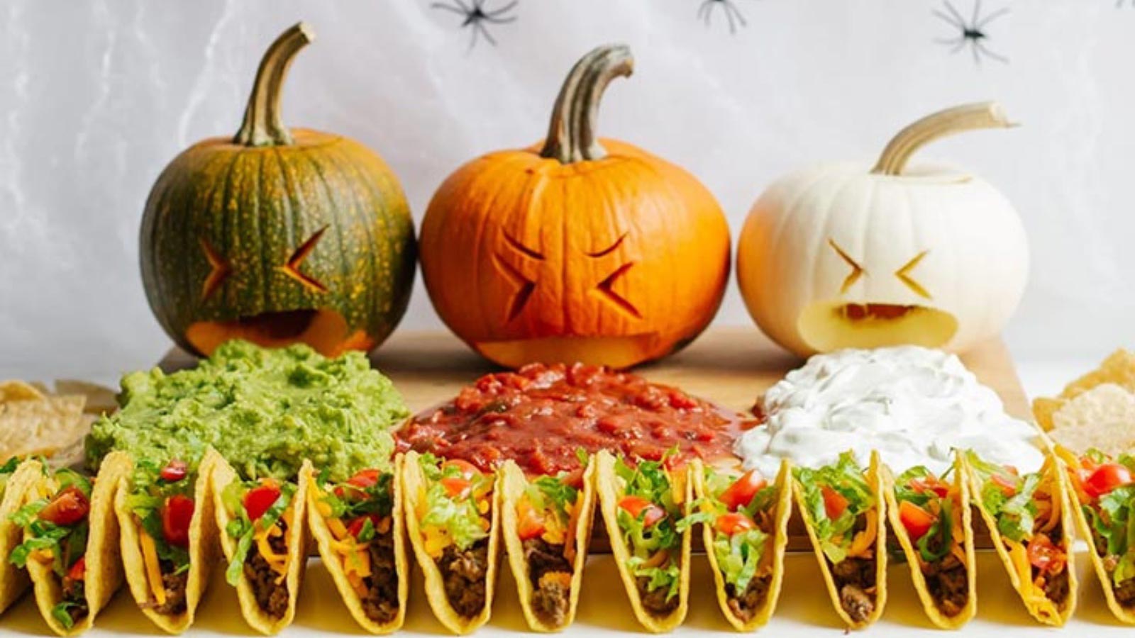 Three pumpkins lined up with faces carved into them to make them look like they are puking. Dip lays in front of them with tacos in front of that.