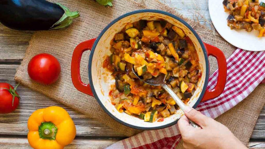 A spoon scoops up a serving of ratatouille from a red pot.