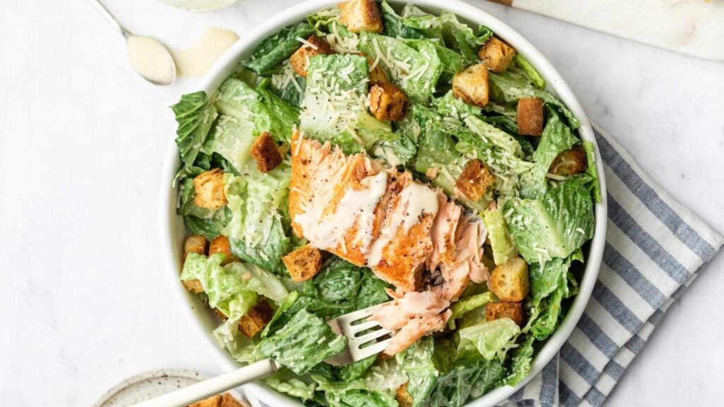 An overhead view of a white ceramic bowl filled with caesar salad and topped with salmon filet.