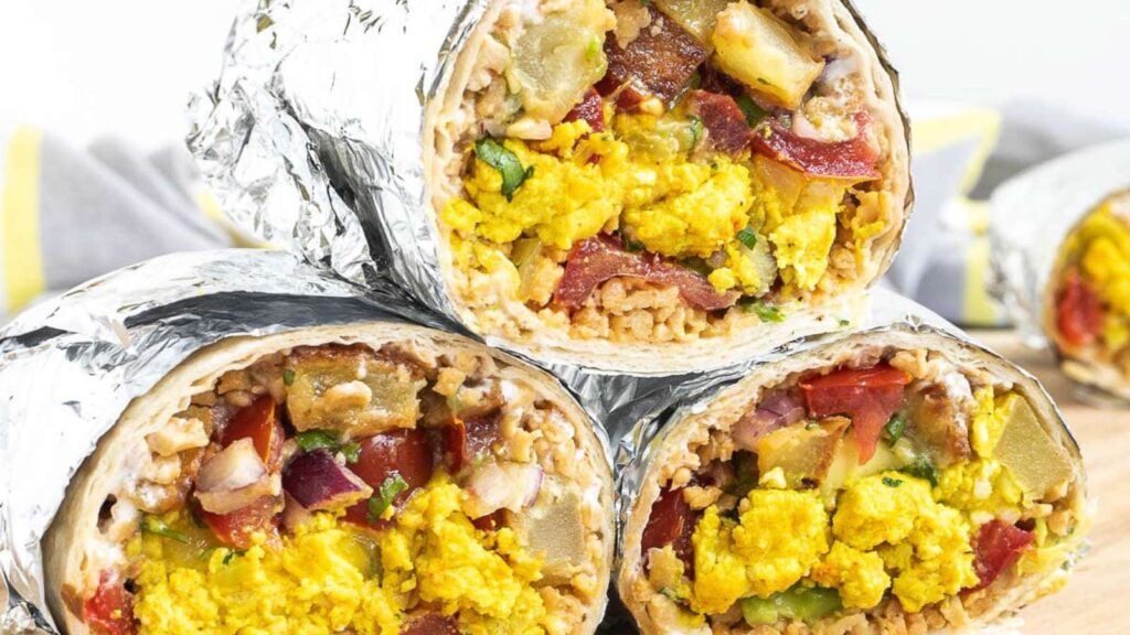Three cut breakfast burritos, wrapped in foil, stacked on a cutting board.