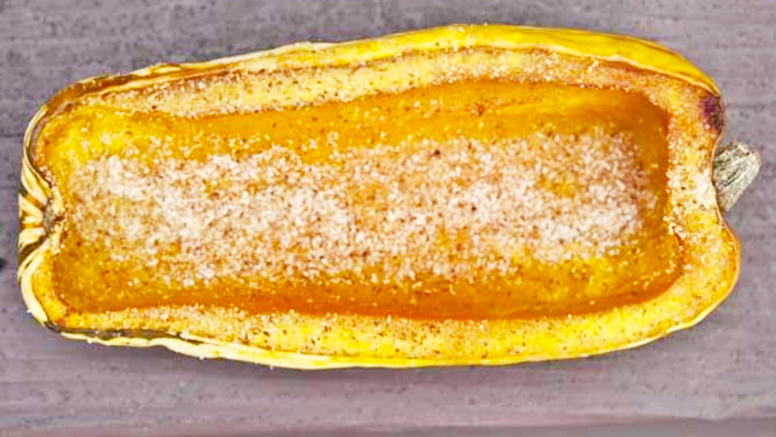 Half a baked delicata squash on a wood surface, sprinkled with spices.