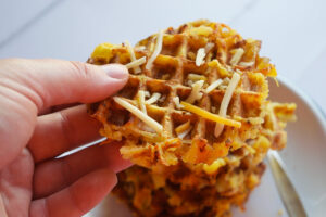 A hand holds a waffle iron smashed potato sprinkled with cheese.