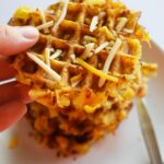 A hand pickup up a smashed potatoes waffle from a stack of them on a white plate.