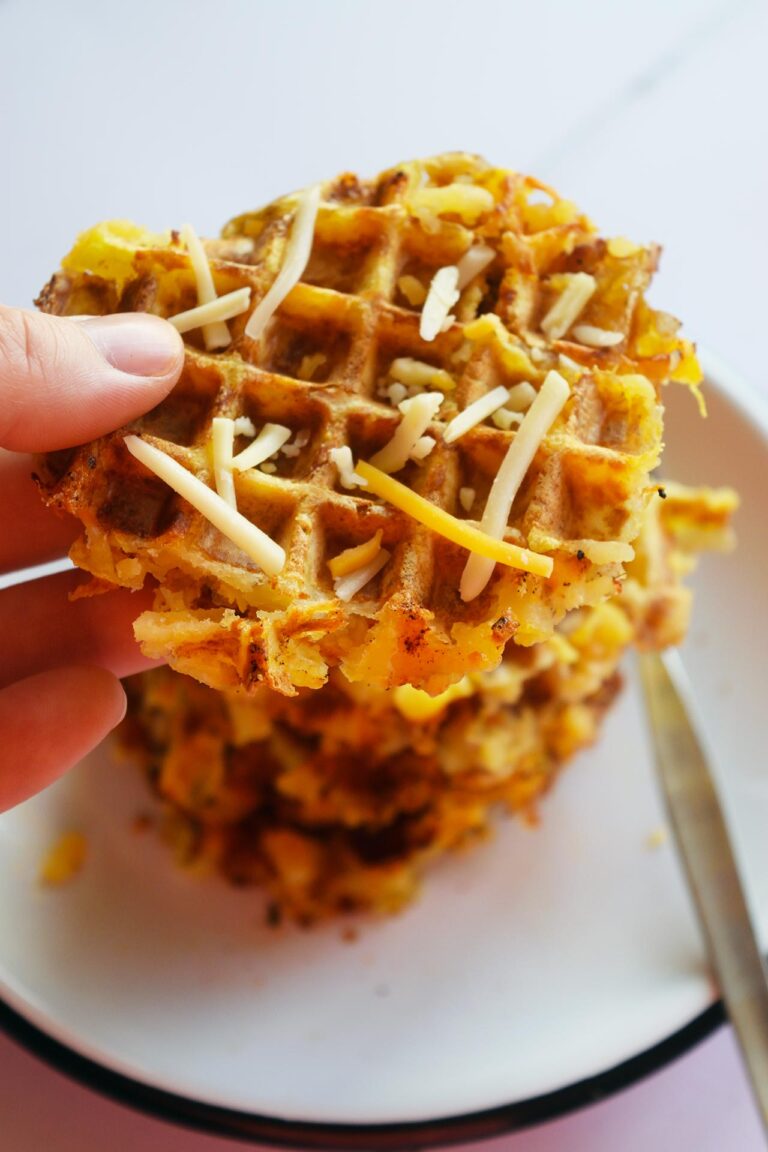 A hand pickup up a smashed potatoes waffle from a stack of them on a white plate.