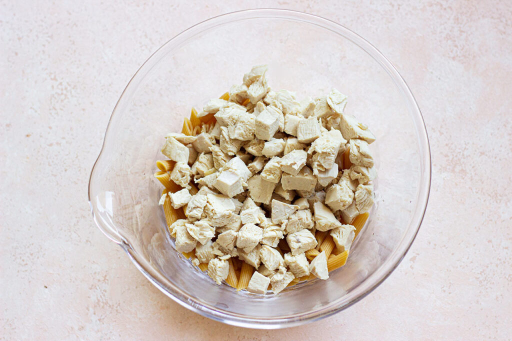 Cooked, chopped chicken added to pasta in a glass mixing bowl.