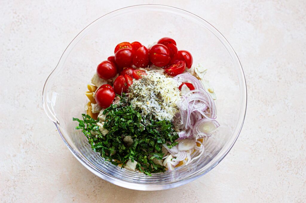 Onions, tomatoes, and basil added to penne pasta in a glass mixing bowl.