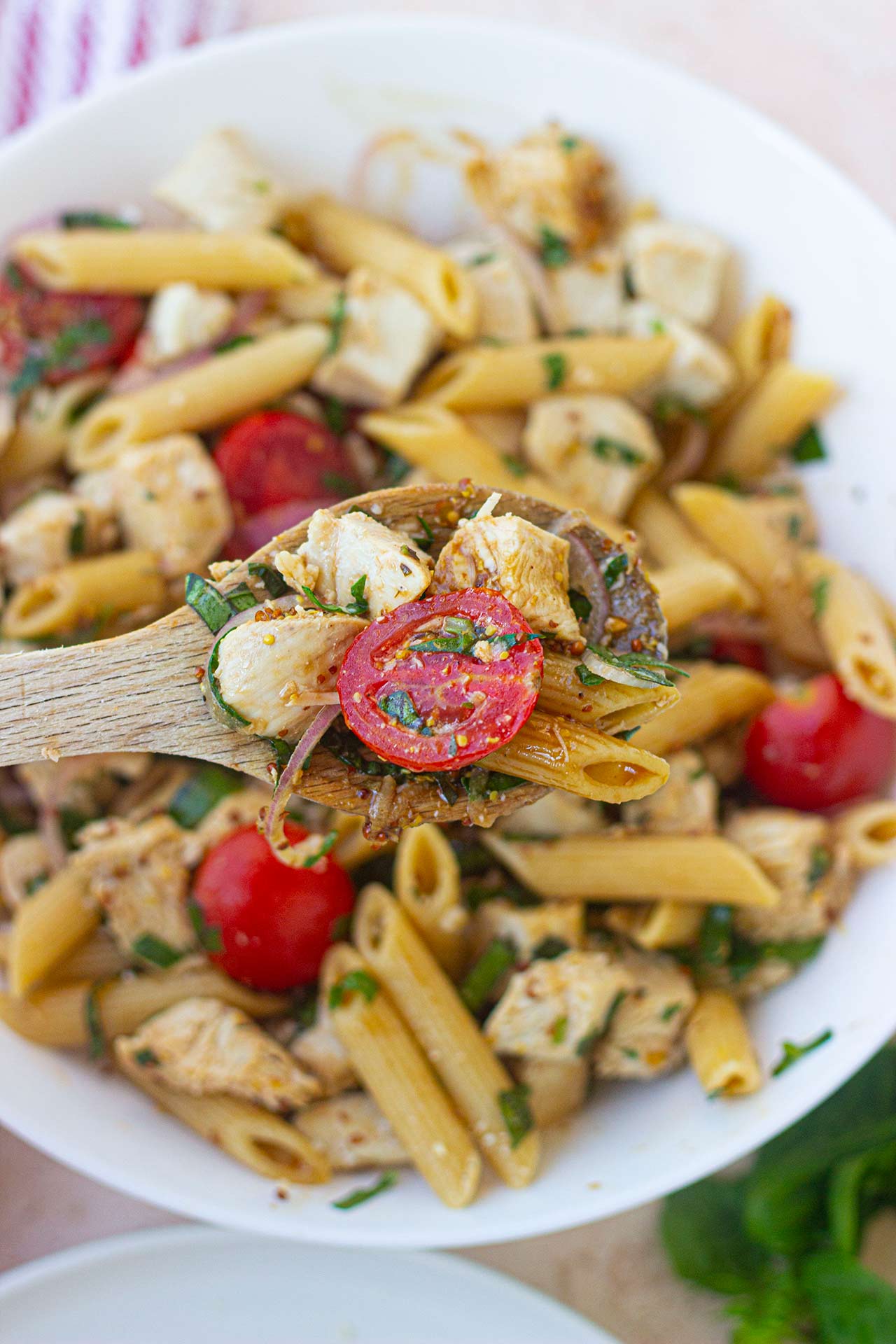 Overhead view. A wood spoon holds a spoonful of chicken pasta salad over a serving bowl filled with it.