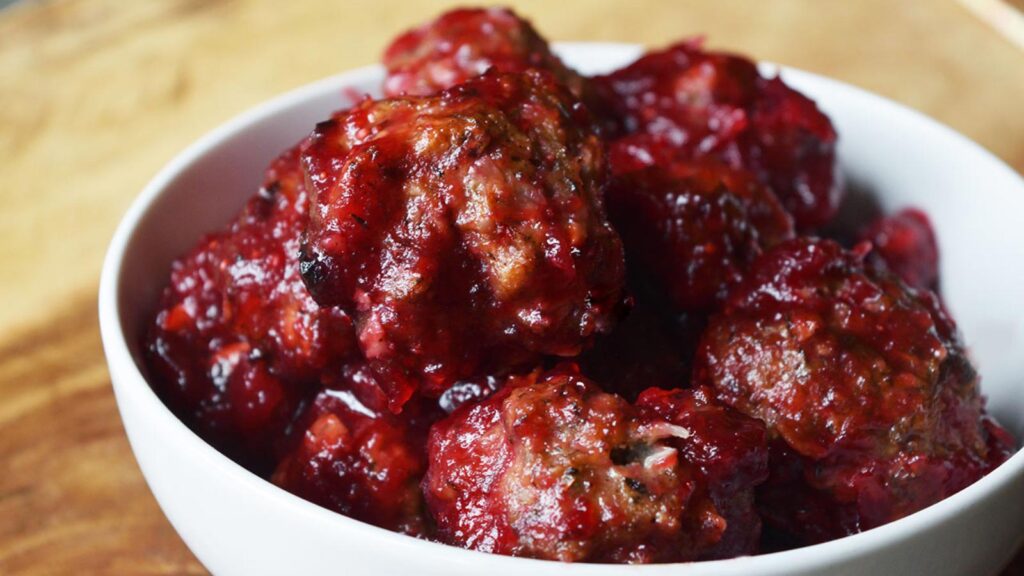 A white bowl filled with cranberry glazed turkey meatballs on a wood surface.
