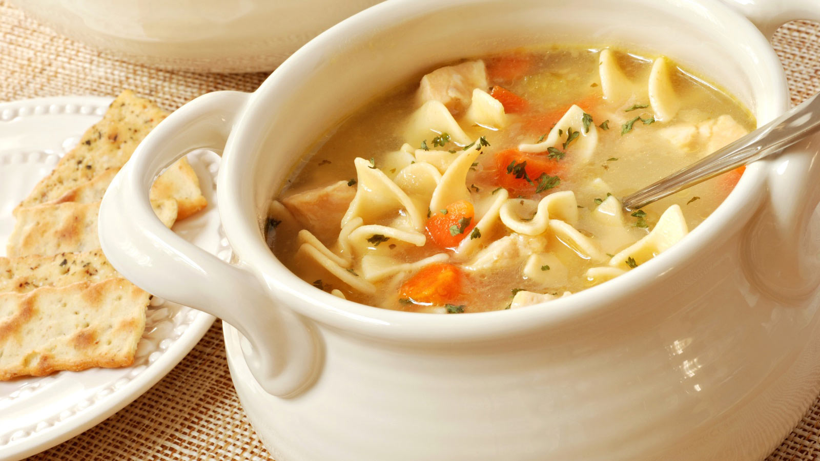Chicken noodle soup in cream colored ceramic bowl with handles. Plate of crackers and soup tureen in background.