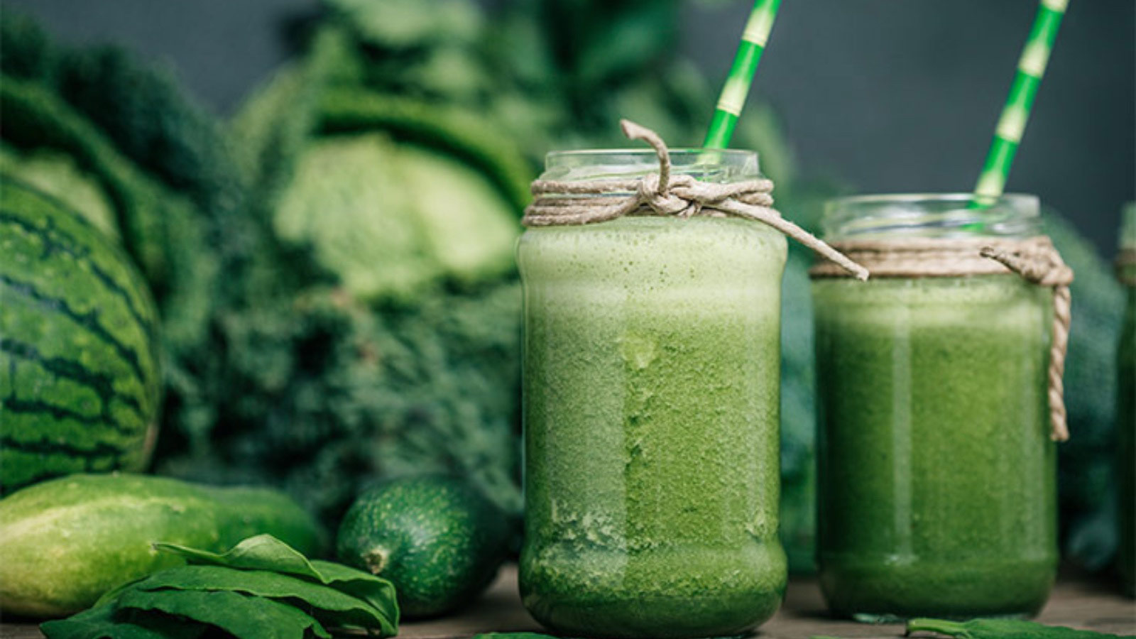Two glasses filled with a green smoothie sit on a wooden surface with lots of green veggies sitting behind the glasses.