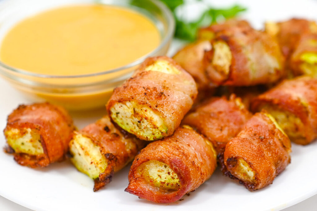 A white plate holds a pile of Bacon-Wrapped Zucchini with a bowl of dip.
