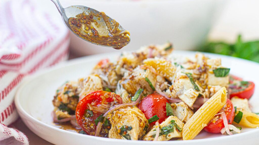 A spoon ladles dressing over a bowl of chicken pasta salad.