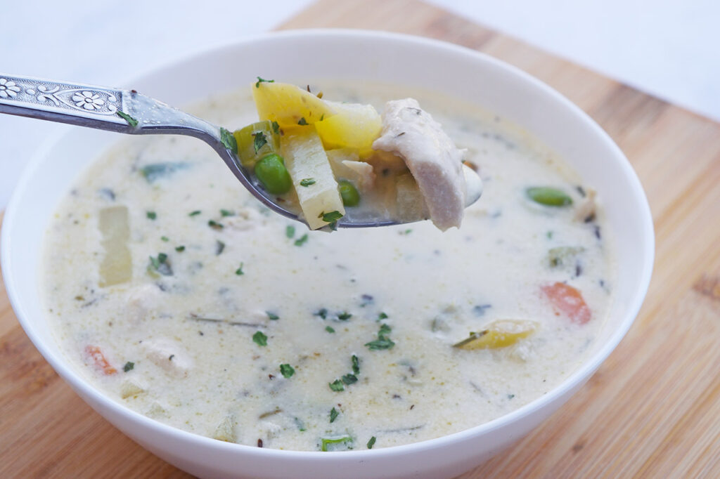 Side view of a white bowl filled with Chicken Pot Pie Soup. A spoon lifts a spoonful out of the bowl.