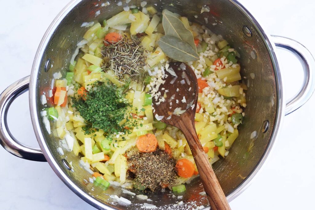 Spices added to a pot of partially cooked veggies. A wooden spoon rests in the pot.
