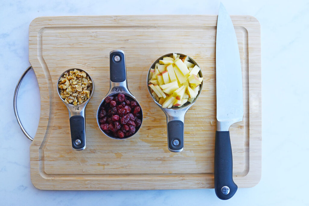 Chopped apples and walnuts as well as dried cranberries in measuring cups sitting on a wood cutting board next to a knife.