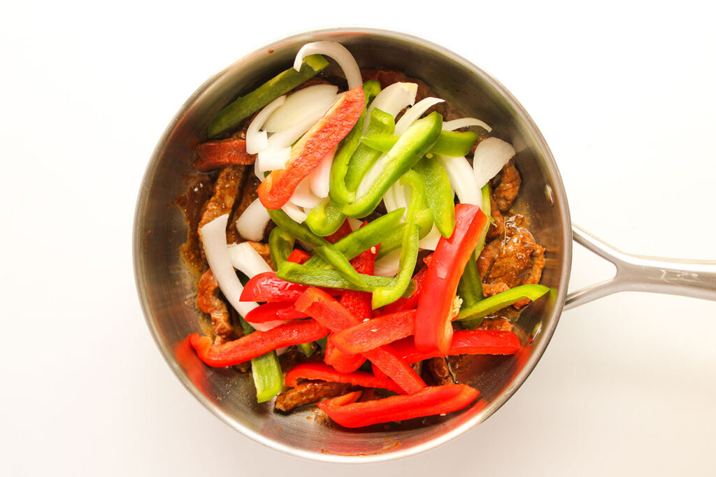 Cut strips of onion and bell peppers added to a skillet with steak strips.