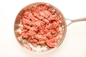 Raw ground beef added to a skillet with sautéed onions.
