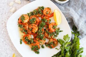 Overhead view of a white, square plate filled with Chimichurri Shrimp and a lemon wedge.