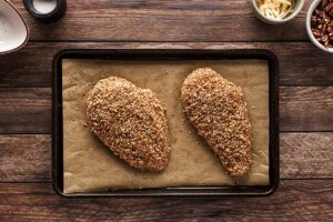 Breaded chicken breasts laying on a parchment-lined baking pan, ready for baking.