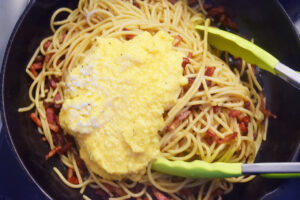 Carbonara sauce added to pasta and bacon bits in as skillet.