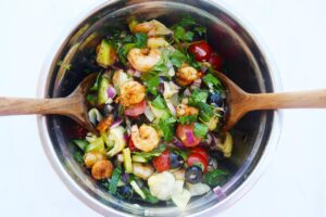 Overhead view of Mediterranean Shrimp Salad in a metal mixing bowl with two wooden spoons. White background.