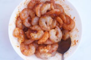 Seasoned shrimp in a white bowl with a metal spoon. White background.