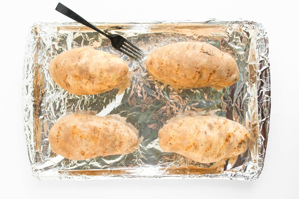 Four Russet potatoes on a foil-lined baking pan with holes poked into them by a fork which rests on the side of the pan.