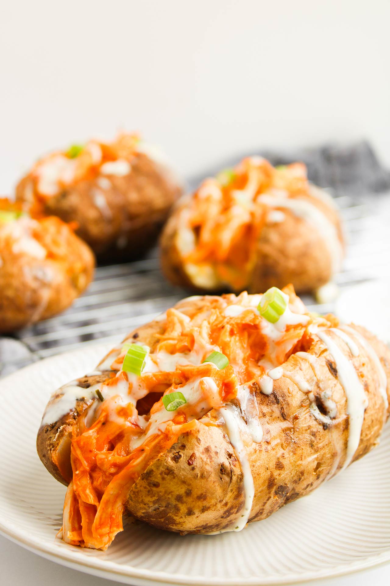 Closeup of a single Buffalo Chicken Stuffed Baked Potato on a white plate. More potatoes sit behind it on a wire rack.