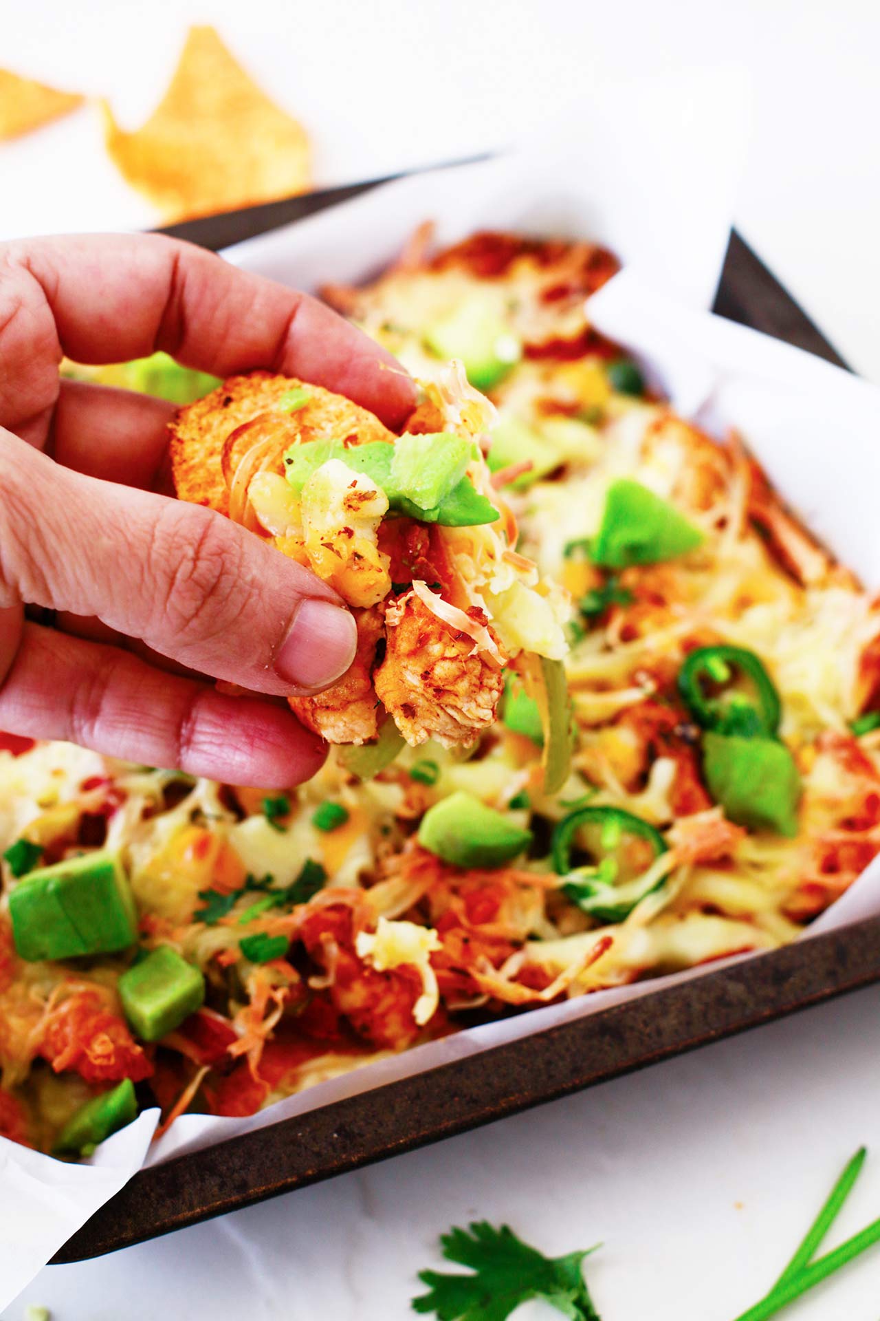 A hand holds a chip with toppings from a pan of Chicken Fajita Nachos.