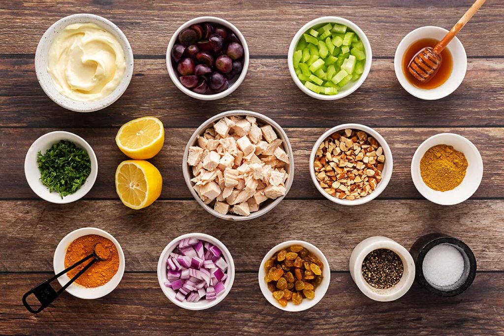 Ingredients for curry chicken salad in individual bowls on a wood surface. Overhead focus.