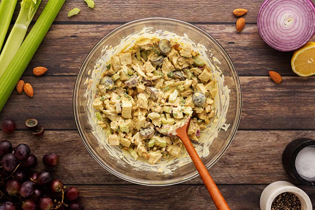 Curry chicken salad mixed with dressing in a mixing bowl with a wood spoon. Overhead focus.