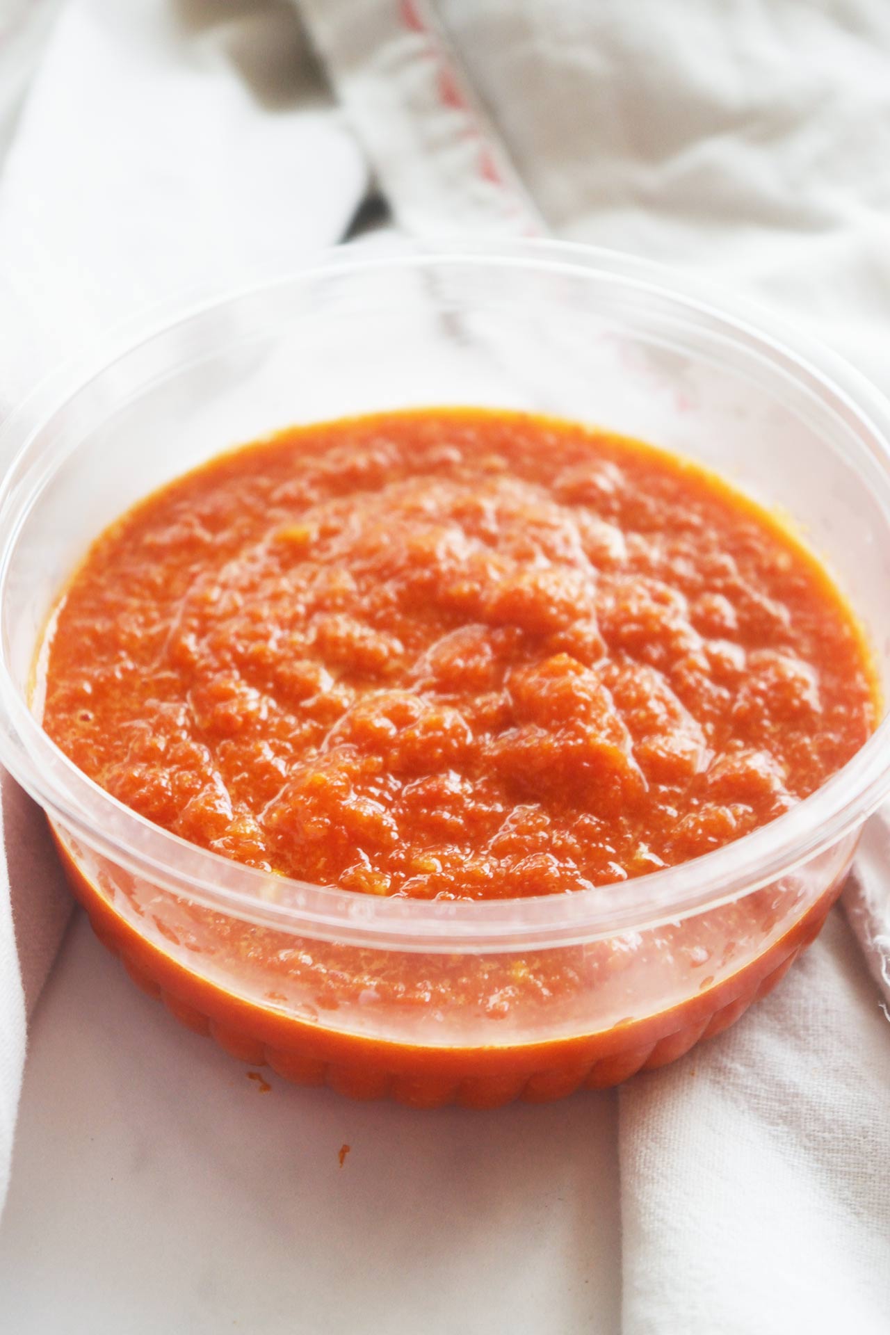 Front view of a small plastic tub of Homemade Sriracha Sauce sitting on a white dish towel.