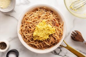 Carbonara mix added to pasta in a skillet with bacon bits and garlic.