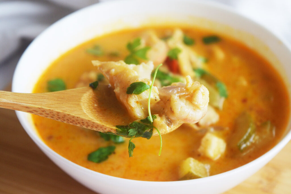 A spoon lifts a bit of this Thai Curry Recipe With Shrimp And Cod out of a white bowl full of it.