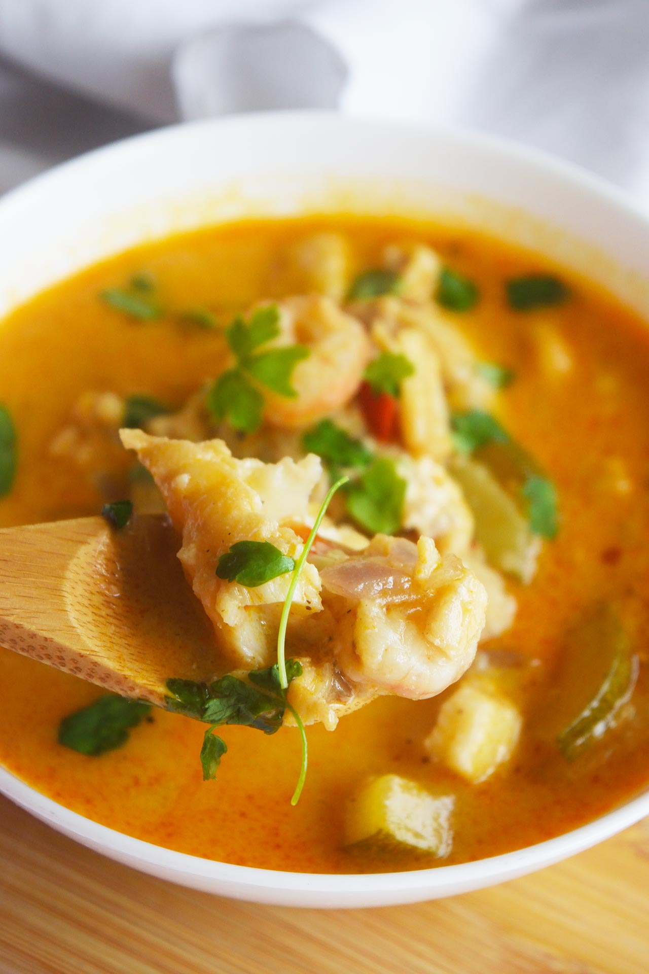 A spoon lifts up a serving of Thai curry with shrimp and cod from a white bowl.