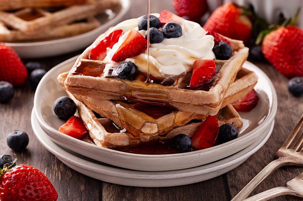 Front view. Pouring syrup over waffled garnished with whipped cream and berries.