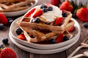 Front view. Pouring syrup over waffled garnished with whipped cream and berries.