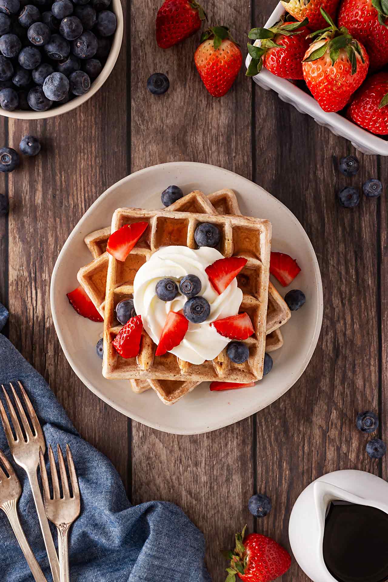 Overhead view of a stack of waffles garnished with whipped cream, blueberries, and cut strawberries.