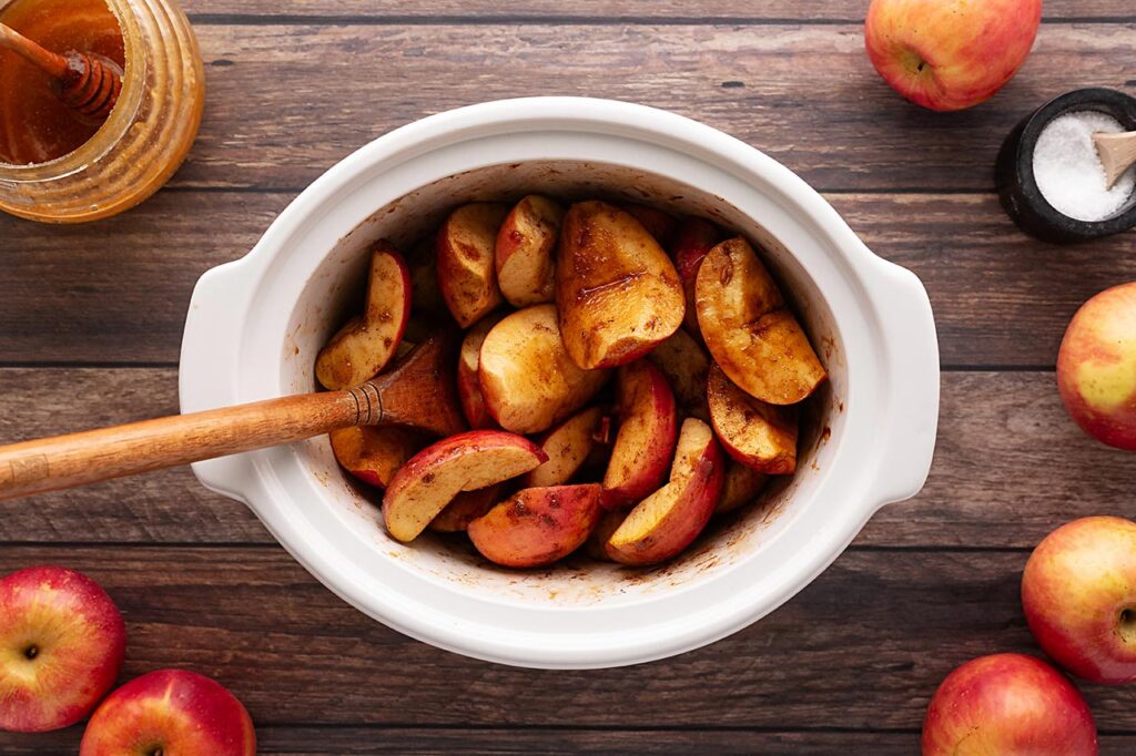 Apples in a white slow cooke crock, stirred until coated with spices and sweeteners.