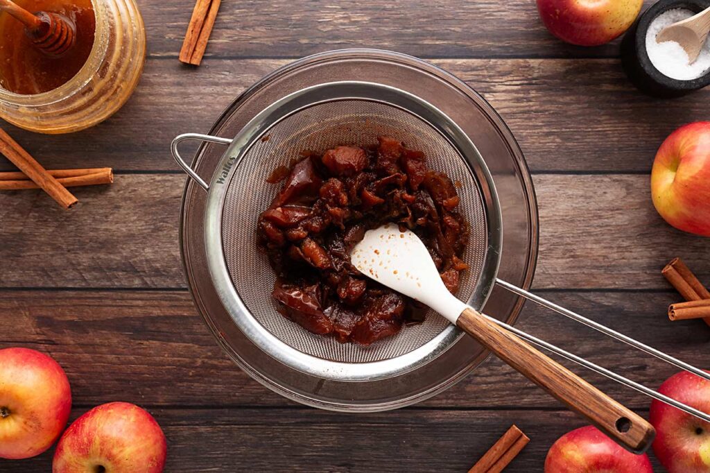 Pressing cooked apples through a fine-meshed sieve over a bowl.