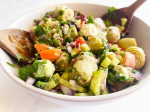 Horizontal orientation. A white bowl filled with Artichoke Salad. Two wooden spoons rest in the bowl