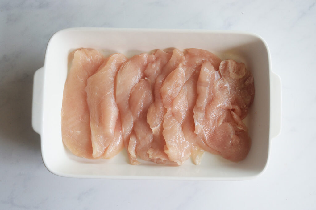 Raw chicken breast slices in a white casserole dish.