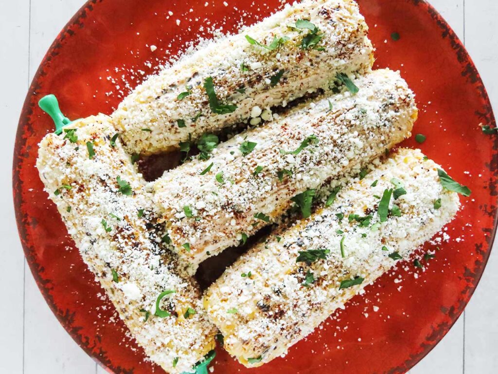 Overhead focus. Corn cobs on a red plate covered in spread, cheese, and green garnish.