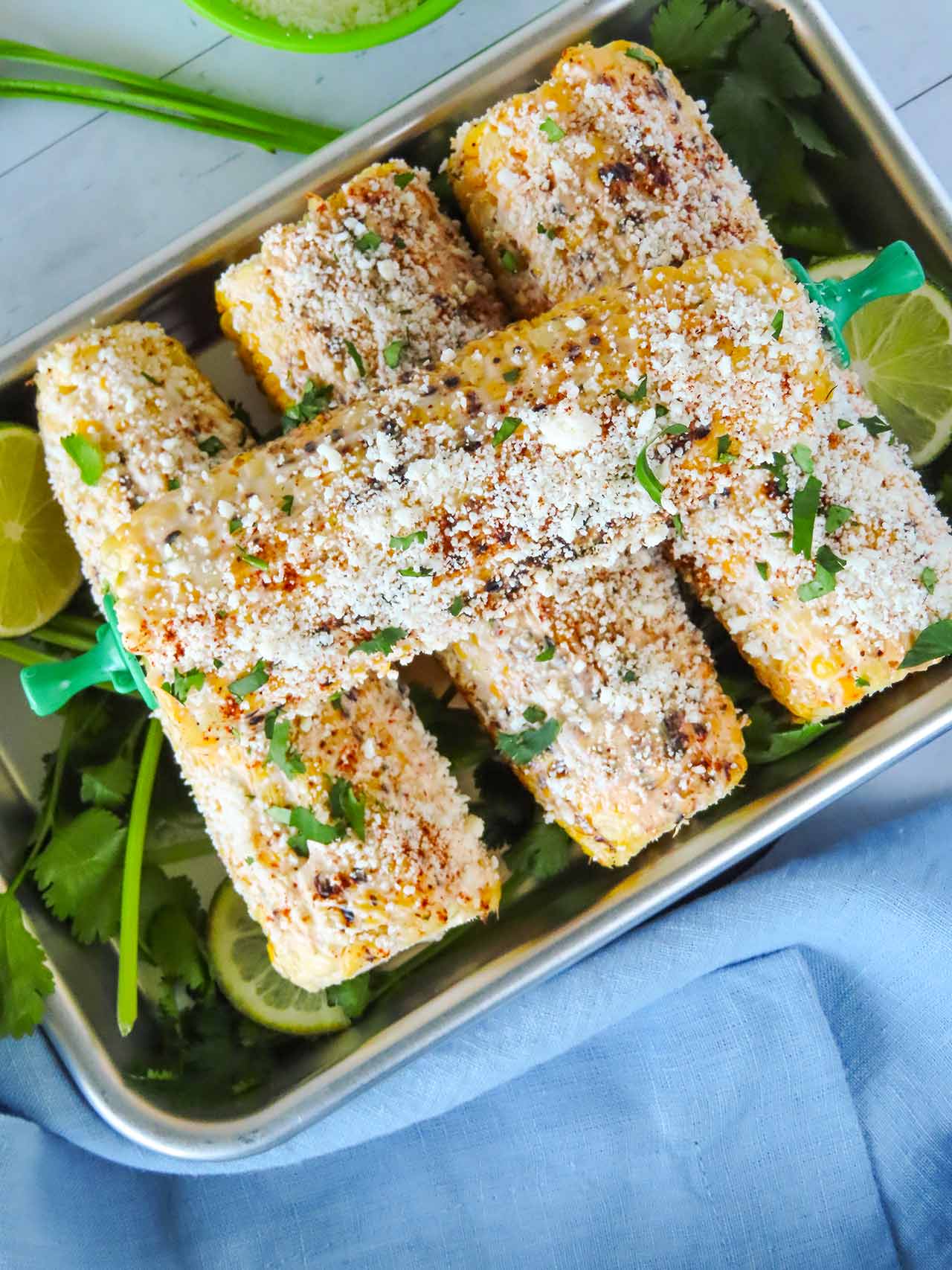 Overhead view of four corn cobs in a metal tray with cilantro and lime slices.