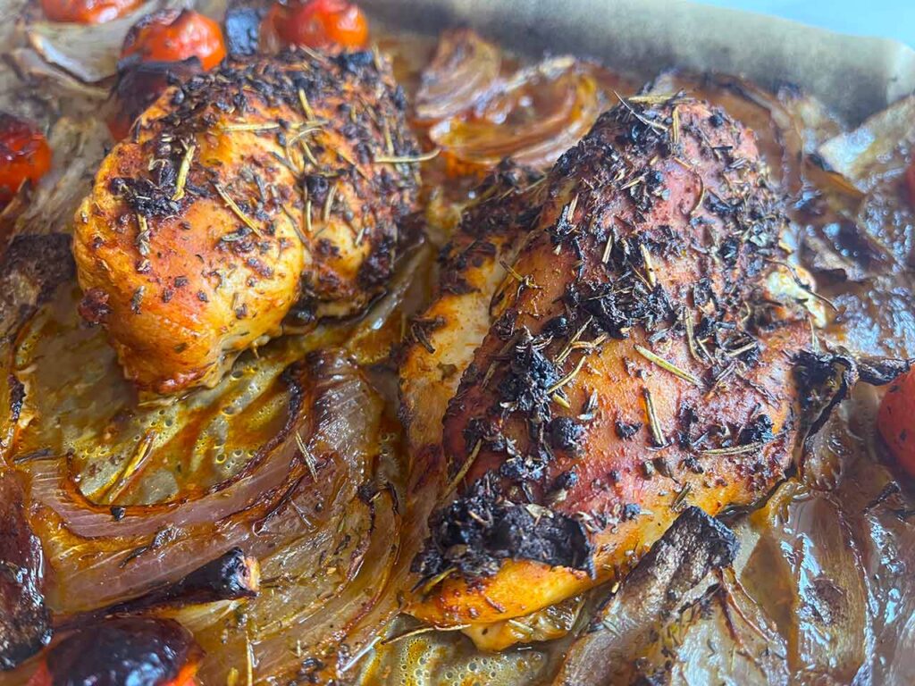 Closeup of two Mediterranean chicken breasts with roasted veggies on a sheet pan.