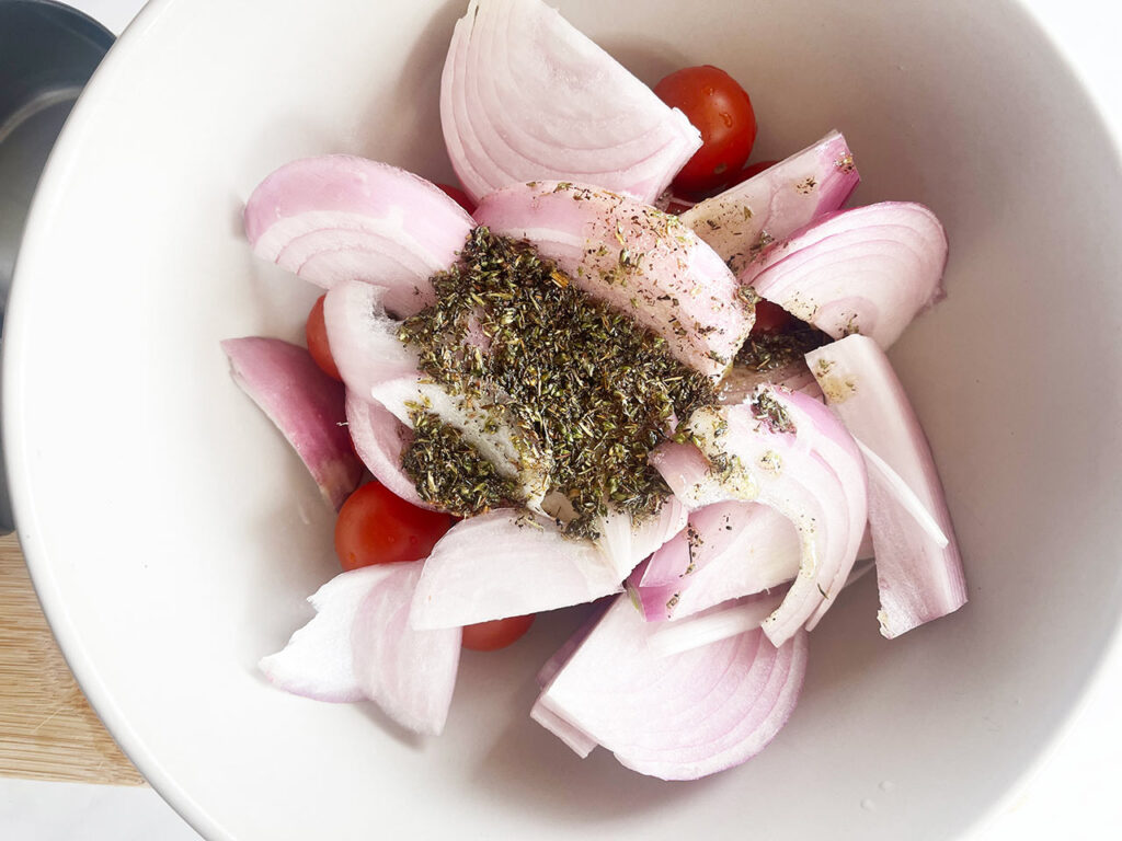 Raw onions and cherry tomatoes in a mixing bowl with dry spices sprinkled on top. Overhead focus.
