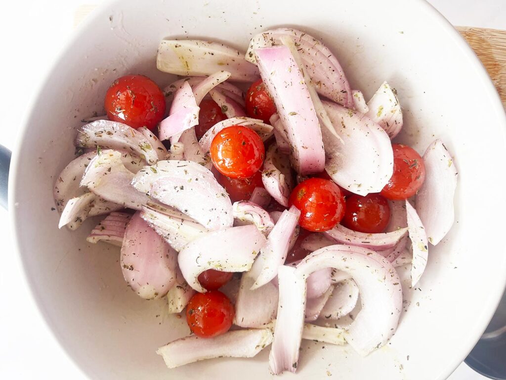 Raw onions and cherry tomatoes tossed with oil and spices in a white mixing bowl.
