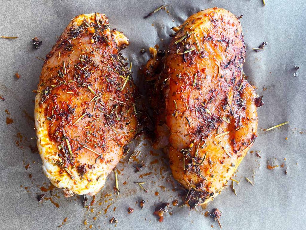 Two, raw, seasoned chicken breasts on a parchment-lined sheet pan. Overhead focus.
